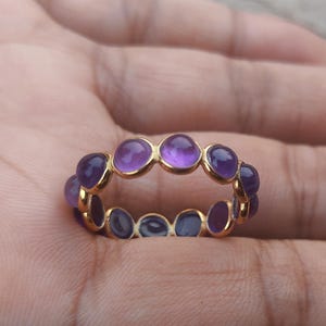 Könnte beinhalten: Ein goldener Ring mit einer Reihe von violetten Edelsteinen. Der Ring ist mit einer Reihe von runden, cabochon geschliffenen Amethyststeinen besetzt.