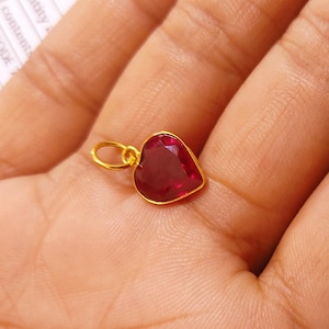 May include: A heart-shaped pendant with a faceted, deep red gemstone set in a gold-colored metal. The pendant has a small loop at the top for a chain. The gemstone is approximately 0.5 inches wide.