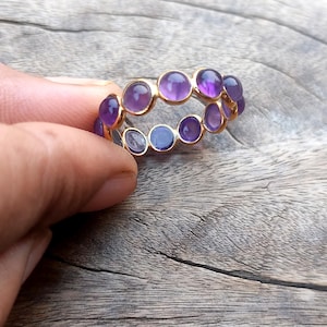May include: A gold ring with a double row of purple amethyst stones. The ring is held in a hand.