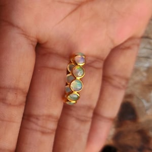 May include: A gold ring featuring a series of oval-shaped opals. The opals display a range of colors, including white, blue, and green. The ring is set against a warm, neutral background.