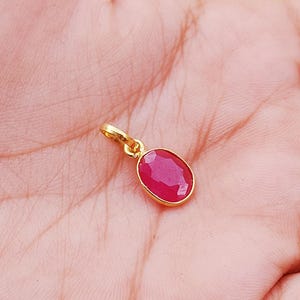 Puede incluir: Un colgante de oro con una piedra preciosa ovalada, facetada, de color rubí. El colgante tiene un diseño sencillo y elegante, con un pequeño lazo para una cadena. El rubí es de un color rojo intenso y vibrante.