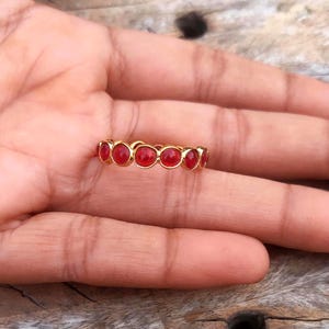 Puede incluir: Un anillo dorado con una serie de gemas rojas vibrantes. Las gemas son de forma ovalada y están engastadas juntas, creando una banda continua de color. El anillo se muestra sobre una superficie de madera.