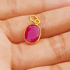 May include: A gold pendant featuring a red oval gemstone. The pendant is resting on a hand.