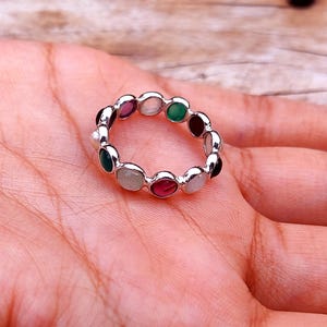 May include: A silver ring with a series of colorful gemstones. The ring features a variety of oval-shaped stones in shades of green, red, purple, and white. The ring is displayed on a person's hand.