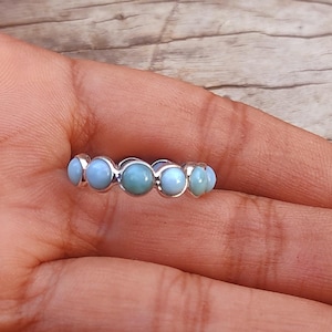 Puede incluir: Un anillo de plata con una fila de piedras preciosas de color azul claro. El anillo está en una mano.