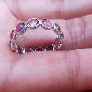 以下が含まれることがあります： 楕円形のピンク色の宝石が連続したパターンでセットされたシルバーリング。 リングは手の上に置かれ、ジュエリーのデザインと色を際立たせています。