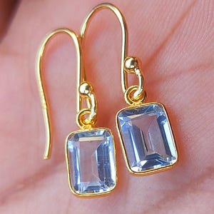 Puede incluir: Un par de pendientes de oro con piedras rectangulares de color azul claro. Los pendientes tienen un diseño simple y elegante.