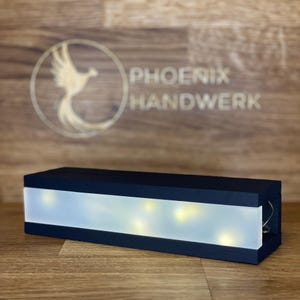 Könnte beinhalten: Eine schwarze Holzkiste mit einer weißen, durchscheinenden Platte, in der sich warmweiße LED-Leuchten befinden. Die Kiste trägt den Text "PHOENIX HANDWERK" auf der Oberfläche dahinter.