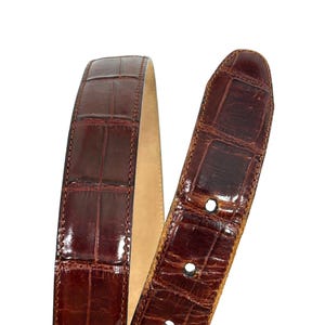 Puede incluir: Primer plano de un cintur&oacute;n de cuero marr&oacute;n con textura de piel de cocodrilo. El cintur&oacute;n tiene un acabado brillante y un interior de color claro. El extremo de la hebilla tiene varios agujeros para ajustar.