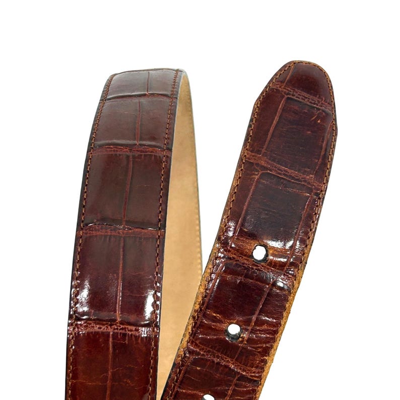 Puede incluir: Primer plano de un cintur&oacute;n de cuero marr&oacute;n con textura de piel de cocodrilo. El cintur&oacute;n tiene un acabado brillante y un interior de color claro. El extremo de la hebilla tiene varios agujeros para ajustar.