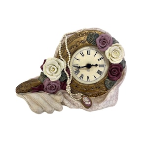 May include: A decorative clock shaped like a hand holding a pearl necklace and roses. The clock face is white with black Roman numerals and a gold trim. The clock is made of ceramic and has a vintage aesthetic.