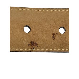 Puede incluir: Primer plano de una secci&oacute;n de un cintur&oacute;n de cuero marr&oacute;n. Se ven dos agujeros circulares, junto con costuras blancas y una mancha oscura. La textura del cintur&oacute;n parece ligeramente desgastada.