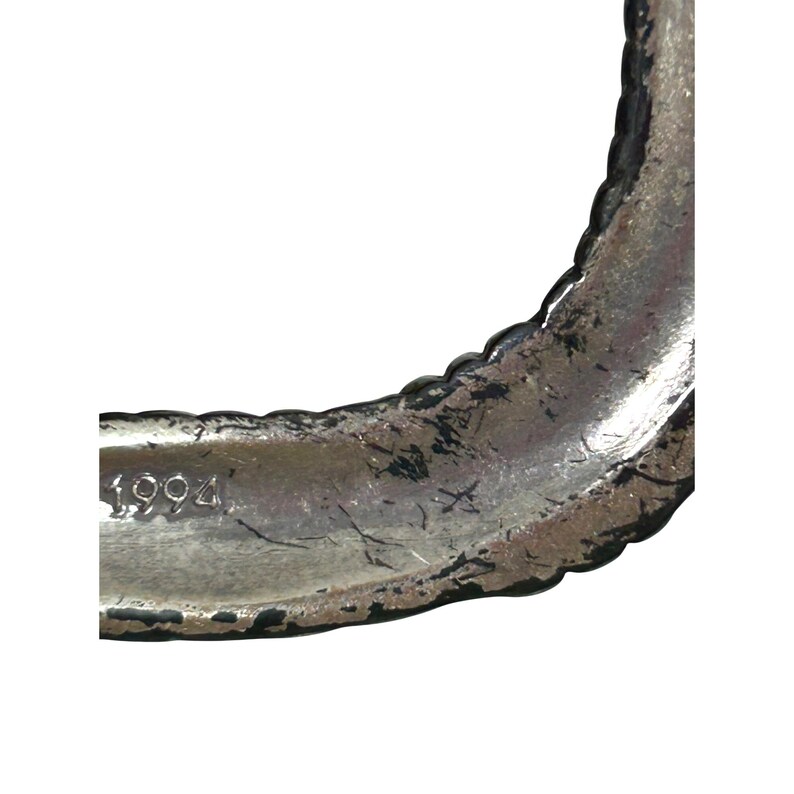 Puede incluir: Primer plano de una pulsera de metal plateado. La pulsera tiene un borde texturizado y festoneado y el n&uacute;mero "1994" es visible. El metal muestra signos de desgaste y envejecimiento.