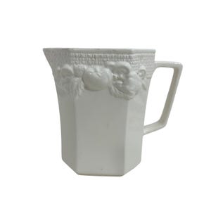 May include: White ceramic pitcher with a geometric shape and a handle. The top edge features a basket weave design with embossed fruit and leaf details. The pitcher is a classic piece for serving beverages.