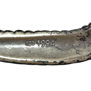 Puede incluir: Primer plano de una pulsera de metal plateado con un borde texturizado. El interior de la pulsera es liso y presenta el s&iacute;mbolo de copyright y el a&ntilde;o 1994.