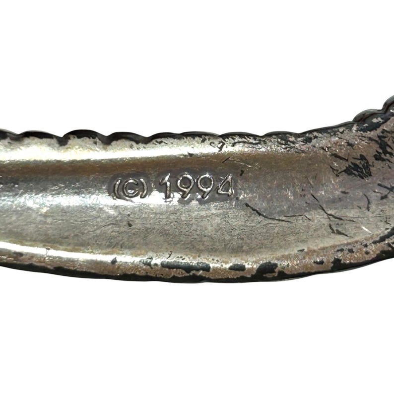 Puede incluir: Primer plano de una pulsera de metal plateado con un borde texturizado. El interior de la pulsera es liso y presenta el s&iacute;mbolo de copyright y el a&ntilde;o 1994.