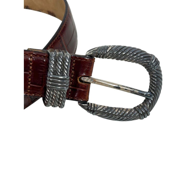 Puede incluir: Un cintur&oacute;n de cuero marr&oacute;n con hebilla plateada. El cintur&oacute;n tiene textura de piel de cocodrilo. La hebilla es de metal plateado con dise&ntilde;o de cuerda trenzada. Un accesorio cl&aacute;sico.