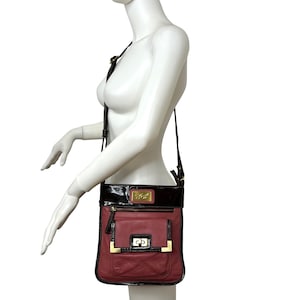 May include: A black and burgundy leather crossbody bag with a gold-tone buckle and a gold logo. The bag has a flap closure and a zippered pocket on the front.