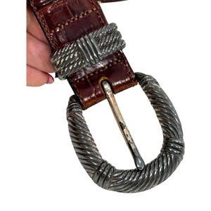 Puede incluir: Un cintur&oacute;n de cuero marr&oacute;n con una hebilla plateada. El cintur&oacute;n tiene un patr&oacute;n texturizado, similar al de un cocodrilo. La hebilla tiene una forma ovalada grande con un dise&ntilde;o de cuerda retorcida. El cintur&oacute;n se abrocha con un pasador de metal plateado.