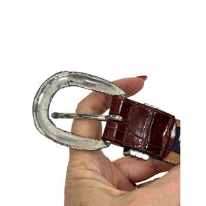 Puede incluir: Un cintur&oacute;n de cuero marr&oacute;n con una hebilla plateada. El cintur&oacute;n tiene una textura de piel de cocodrilo y una hebilla plateada con un borde decorativo. El cintur&oacute;n se sostiene en una mano.