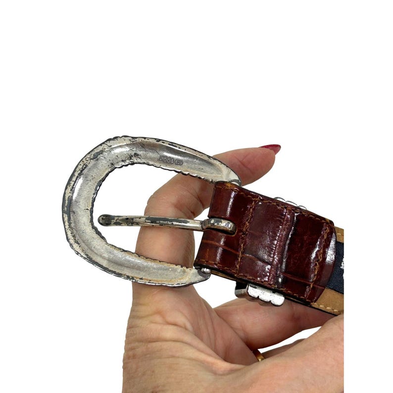 Puede incluir: Un cintur&oacute;n de cuero marr&oacute;n con una hebilla plateada. El cintur&oacute;n tiene una textura de piel de cocodrilo y una hebilla plateada con un borde decorativo. El cintur&oacute;n se sostiene en una mano.