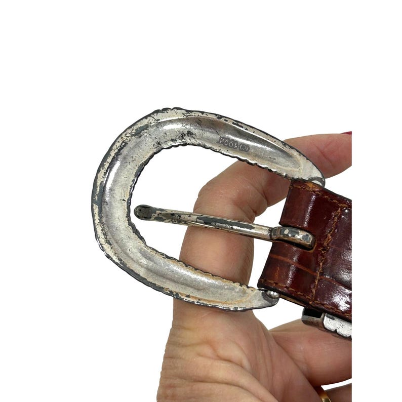 Puede incluir: Primer plano de una hebilla de cintur&oacute;n plateada con un cintur&oacute;n de cuero marr&oacute;n. La hebilla tiene un dise&ntilde;o ovalado texturizado. El cintur&oacute;n de cuero es de color marr&oacute;n intenso con un patr&oacute;n de cocodrilo.