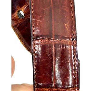 Puede incluir: Primer plano de un cintur&oacute;n de cuero marr&oacute;n con textura de piel de cocodrilo. El cintur&oacute;n tiene forma rectangular con un borde cosido y un agujero para la hebilla. El cuero tiene un acabado brillante.