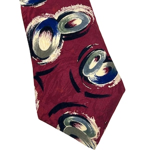 May include: A burgundy tie with an abstract design. The tie features large, overlapping circles in shades of blue, gray, and cream, with black brushstrokes. The tie is made of a smooth fabric.