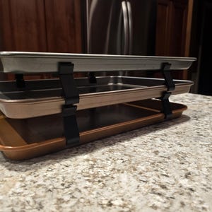 May include: Three baking sheets stacked on top of each other with black metal clips holding them together. The baking sheets are different colors: silver, brown, and copper.