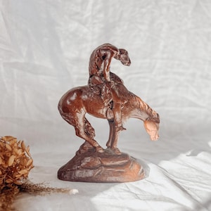 May include: A brown wooden sculpture of a cowboy riding a bucking bronco. The sculpture is detailed, showing the cowboy leaning forward and the horse in mid-air. The base is a rocky formation.