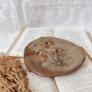 May include: A brown ceramic dish with three pressed flowers. The dish is placed on an open book with visible text. Dried plants are to the left of the dish. The dish is approximately 6 inches in diameter.