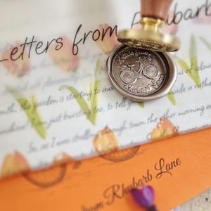 May include: A wax seal stamp with a wooden handle and a brass head featuring a bicycle design, pressing into a metallic gold wax seal on a white envelope with floral patterns and the text Letters from Rhubarb Lane printed on orange paper.