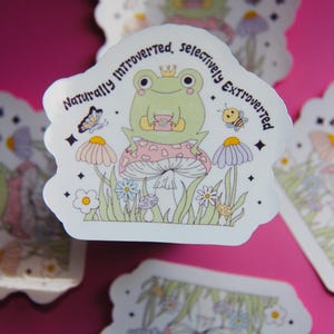 May include: A sticker featuring a cartoon frog wearing a crown and sitting on a mushroom. The sticker has the text "Naturally Introverted, Selectively Extroverted" and includes illustrations of flowers, a butterfly, and a bee.