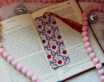 Può includere: Un libro aperto con un segnalibro con un motivo a ciliegie e fiocchi. Perline di legno rosa e sfere a specchio circondano il libro. Il segnalibro ha un nappa rossa. Il libro è aperto al capitolo XXVII.