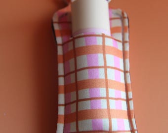 Könnte beinhalten: Ein Lippenbalsamhalter mit einem Metall-Schlüsselring. Der Halter besteht aus Stoff mit einem karierten Muster in Orange, Rosa und Weiß. Ein zylindrischer Lippenbalsam ist teilweise sichtbar.