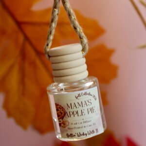 Pode incluir: Um pequeno frasco difusor de carro em vidro transparente com tampa de madeira clara e corda trançada. O rótulo apresenta um gráfico de tarte de maçã e o texto Fall Collection 2024, MAMA'S APPLE PIE, 8 ml Car Diffuser, Hand Poured in Cats NY, Jellin' Wicky With It.