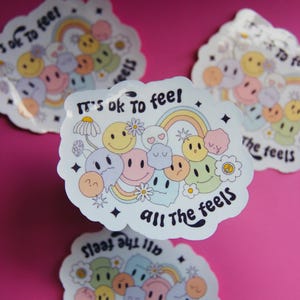 Könnte beinhalten: Mehrere Aufkleber mit dem Text „It's ok to feel all the feels“. Die Aufkleber zeigen eine Reihe von lächelnden und traurigen Gesichtern, Regenbögen und Blumen in Pastellfarben. Die Aufkleber befinden sich auf einem rosa Hintergrund.