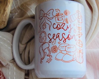 May include: White ceramic mug with a large handle, featuring a red-orange illustration of cozy season elements. The design includes a bow, jar of honey, scarf, headphones, hat, boots, and the words "cozy season".