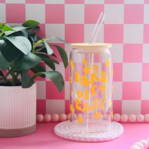 May include: A clear glass tumbler with a bamboo lid and straw, featuring the text "TAKE IT EASY" in yellow. The tumbler sits on a white crocheted coaster. A potted plant and pink checkered background complete the scene.