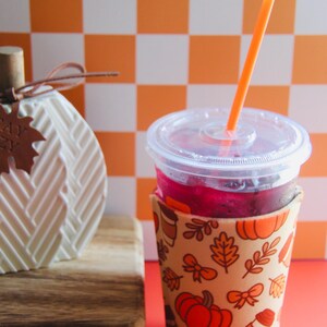 Puede incluir: Un vaso de plástico transparente con una bebida roja y una pajita naranja se encuentra en un portavasos estampado. El portavasos presenta calabazas, hojas y tazas de café. Una decoración de calabaza blanca y una tabla de madera están en el fondo.