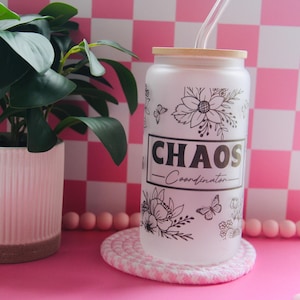 May include: A white glass can-shaped tumbler with a wooden lid and a clear straw. The tumbler features black floral and butterfly designs, with the word "CHAOS" and "-Coordination-" printed on it. It sits on a pink and white coaster, next to a potted plant.