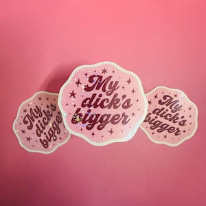 Könnte beinhalten: Drei rosa Aufkleber mit gewelltem Rand und dem Text "My dick's bigger" in einer Schreibschrift. Die Aufkleber haben ein Stern- und Glitzerdesign und sind vor einem rosa Hintergrund angeordnet.