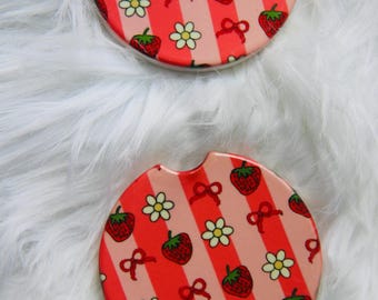 May include: Two round car coasters with a strawberry and floral pattern. The coasters feature a red and pink striped background with strawberries, white flowers, and red bows. Each coaster has a small notch for easy removal.