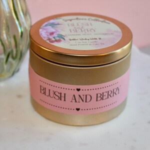 Może przedstawiać: Złota metalowa puszka na świecę z różową etykietą z napisem BLUSH AND BERRY. Wieczko posiada okrągłą etykietę z motywami kwiatowymi oraz tekstem BLUSH AND BERRY, 7 oz Soy Candle, Hand Poured In Ohio NY. Puszka ma cylindryczny kształt.