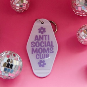 以下が含まれることがあります： 「ANTI SOCIAL MOMS CLUB」の文字が紫で書かれた、ライラック色のモーテル風キーホルダー。キーホルダーは花のグラフィックで装飾され、ピンクの背景に小さなミラーボールが配置されています。
