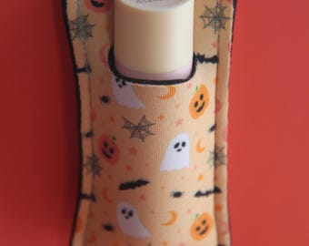Könnte beinhalten: Ein Halloween-Lippenbalsamhalter mit Schlüsselanhänger. Der Halter ist hellorange und hat ein Muster aus Geistern, Kürbissen und Spinnweben. Der Lippenbalsam ist hellbeige. Der Halter ist schwarz eingefasst.