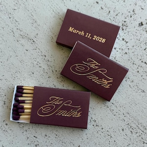 May include: Three rectangular matchboxes in deep burgundy with gold foil lettering. Two boxes are closed, displaying the text "The Smiths" in script and "March 11, 2028" in print. One box is open, revealing wooden matches with dark purple tips.