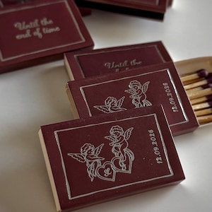 May include: Several rectangular matchboxes in deep burgundy with silver foil detailing. The boxes feature illustrations of two cherubs, hearts, the text "Until the end of time," and the date "12.09.2026." One box is open, revealing wooden matches.