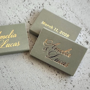 May include: Three rectangular sage green boxes with gold foil lettering. Two boxes feature the names Amelia & Lucas in a cursive script, while the third box displays the date March 11, 2028, in a clean, sans-serif font on the top surface.