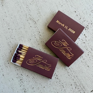 May include: Three rectangular matchboxes in a deep burgundy color with gold foil lettering. Two boxes feature the script text The Smiths, while one box displays the date March 11, 2028. One box is partially open, revealing wooden matches with dark purple tips.